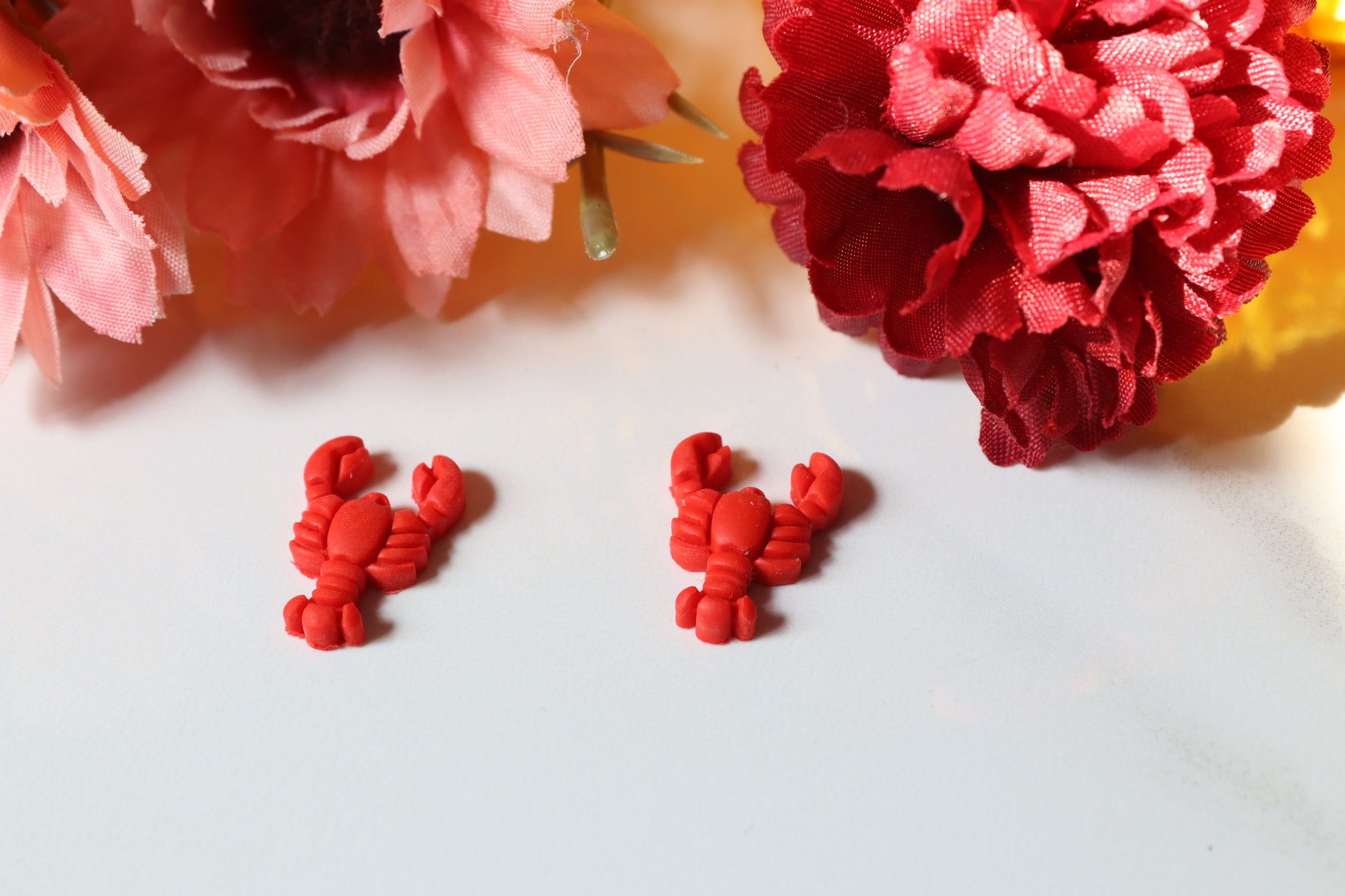 Red lobster-shaped earrings on a white surface with pink and red flowers in the background.