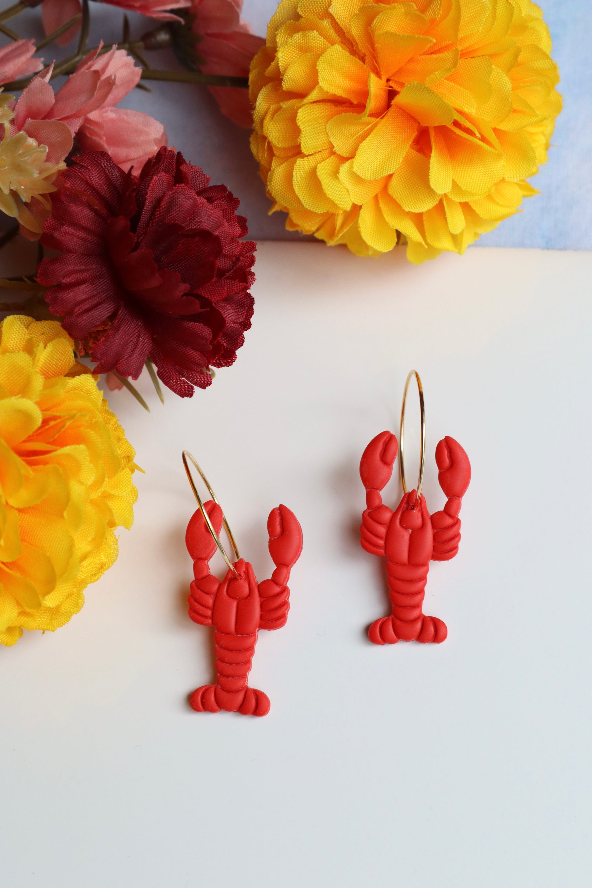 Red lobster-shaped earrings on a white surface with yellow flowers in the background